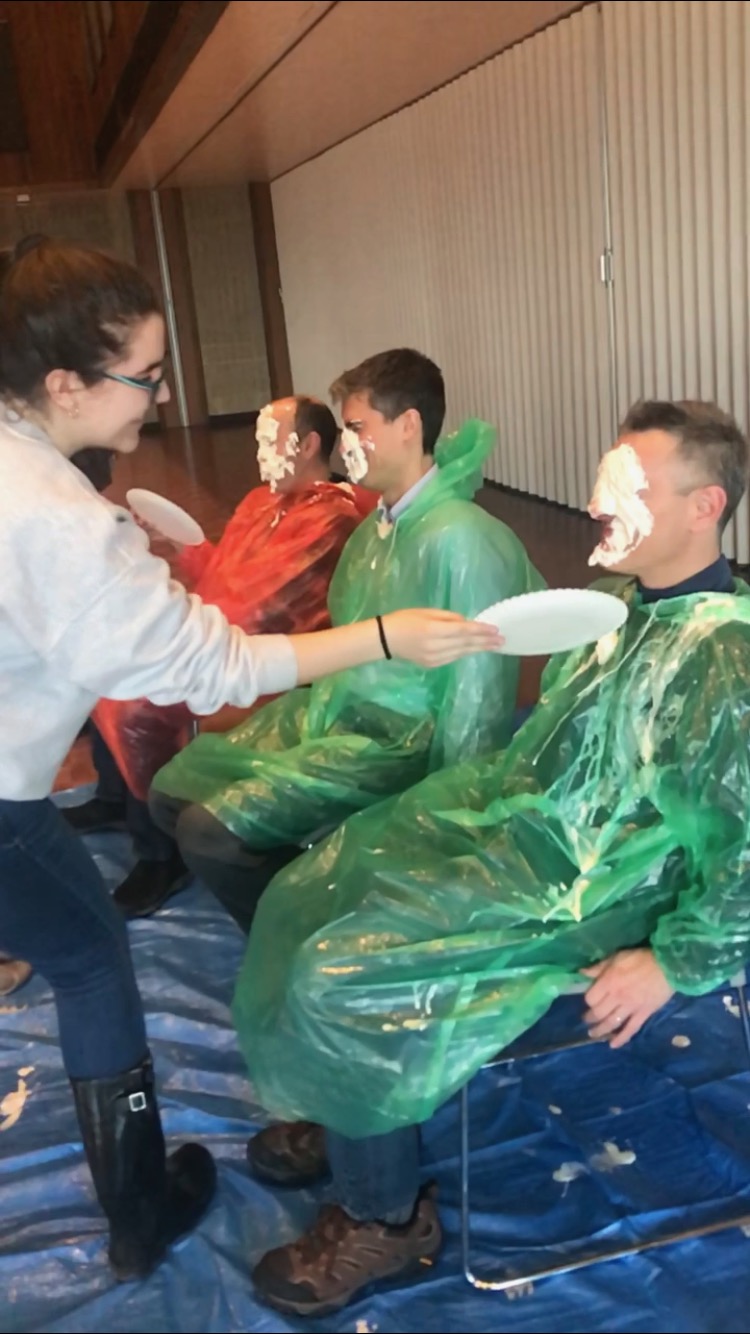 A girl pies a man in the face. Two other men in the background have whipped cream on their faces.
